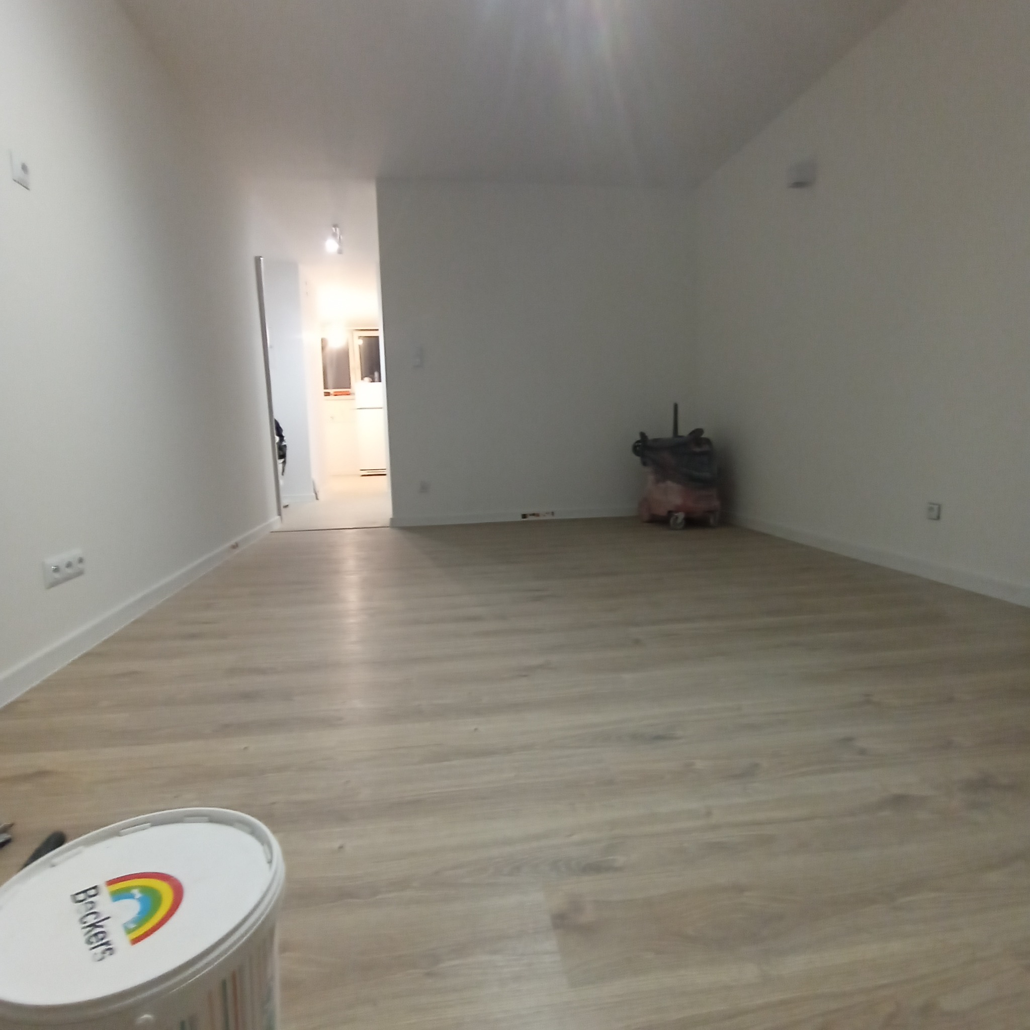 Pomalowane na biało wnętrze mieszkania z jasną podłogą z paneli, widoczny pojemnik z farbą Beckers i narzędzia malarskie oraz odległa kuchnia.