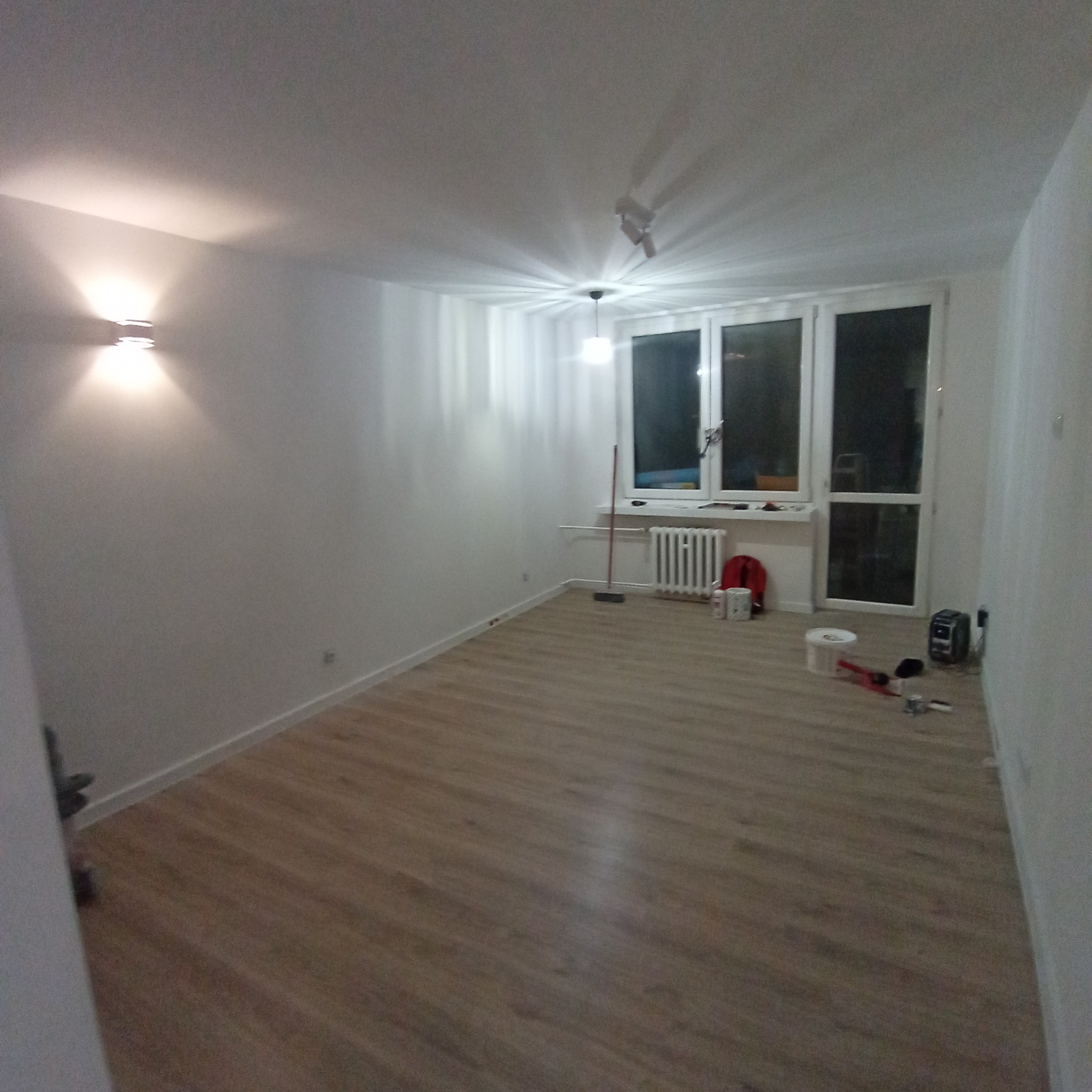 Pusty pokój po odświeżeniu, z panelami podłogowymi, pomalowanymi ścianami na biało, widoczne narzędzia malarskie i sprzęt grzewczy pod oknem.