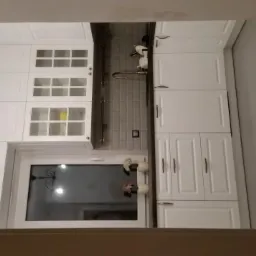 Biała kuchnia z frezowanymi frontami, szarym blatem i backsplashiem z płytek typu metro, oświetlona dekoracyjnym, kutym kinkietem.