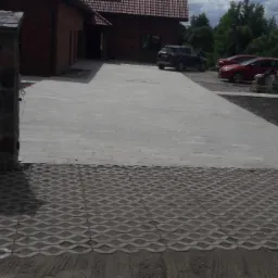 Podjazd do domu wyłożony jasnoszarą kostką brukową, przechodzący w ażurową kostkę trawnikową, z bramą wjazdową z kamiennymi słupkami i czarnym metalowym ogrodzeniem.