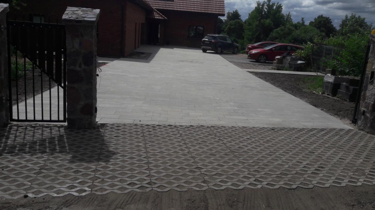 Podjazd do domu wyłożony jasnoszarą kostką brukową, przechodzący w ażurową kostkę trawnikową, z bramą wjazdową z kamiennymi słupkami i czarnym metalowym ogrodzeniem.