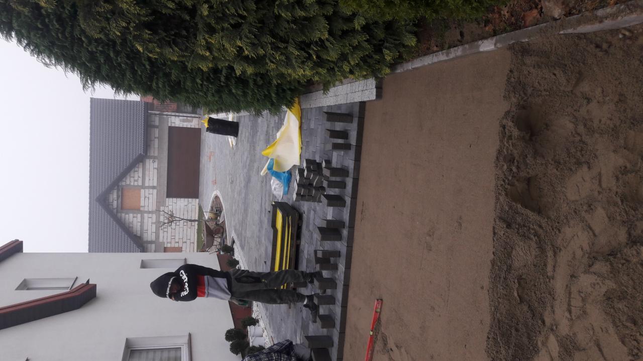 Ciemnoszara kostka brukowa układana na podsypce piaskowej, widoczny poziomica, częściowo ułożona nawierzchnia, w tle dom jednorodzinny z garażem.