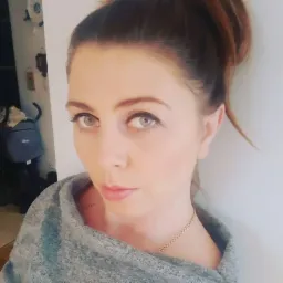 Portret kobiety z włosami upiętymi w kok, ubranej w szary sweter i złotą biżuterię. Uwagę przykuwają jej jasne oczy i delikatny makijaż, a w tle widoczny jest plecak.