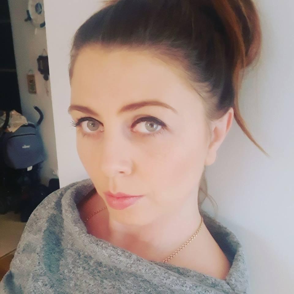 Portret kobiety z włosami upiętymi w kok, ubranej w szary sweter i złotą biżuterię. Uwagę przykuwają jej jasne oczy i delikatny makijaż, a w tle widoczny jest plecak.