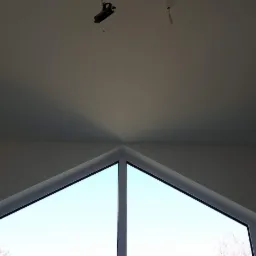 Białe, trójkątne okno na poddaszu z widokiem na niebo, sufit z widocznymi kablami i elementami oświetleniowymi.