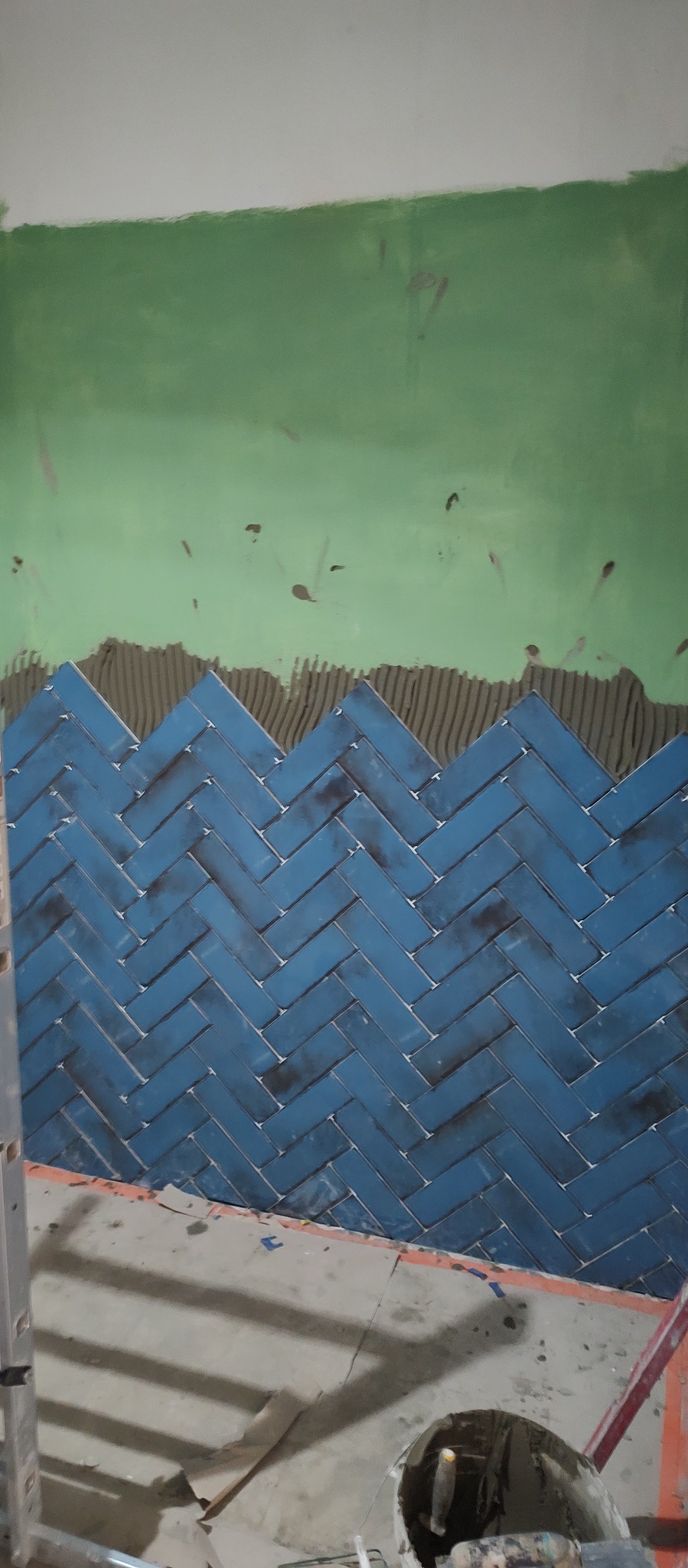 Niebieska glazura ułożona w jodełkę na ścianie łazienki, widoczny fragment zielonej ściany powyżej i zaprawa klejowa, etap prac wykończeniowych.