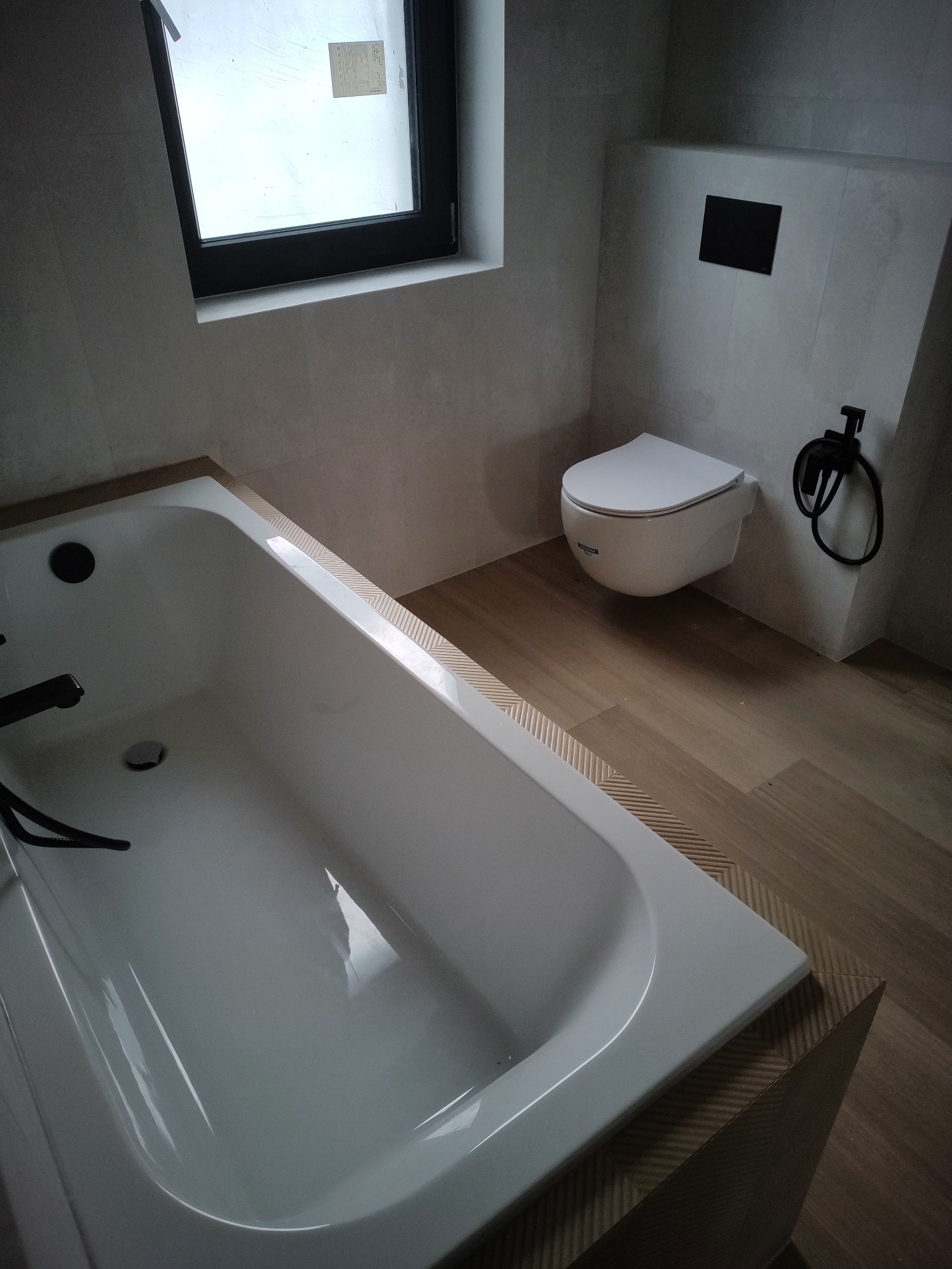 Nowoczesna łazienka w trakcie wykańczania, z białą wanną, podwieszaną toaletą i drewnopodobną podłogą, widok z góry na wannę, z czarnymi akcentami armatury i ramą okienną.