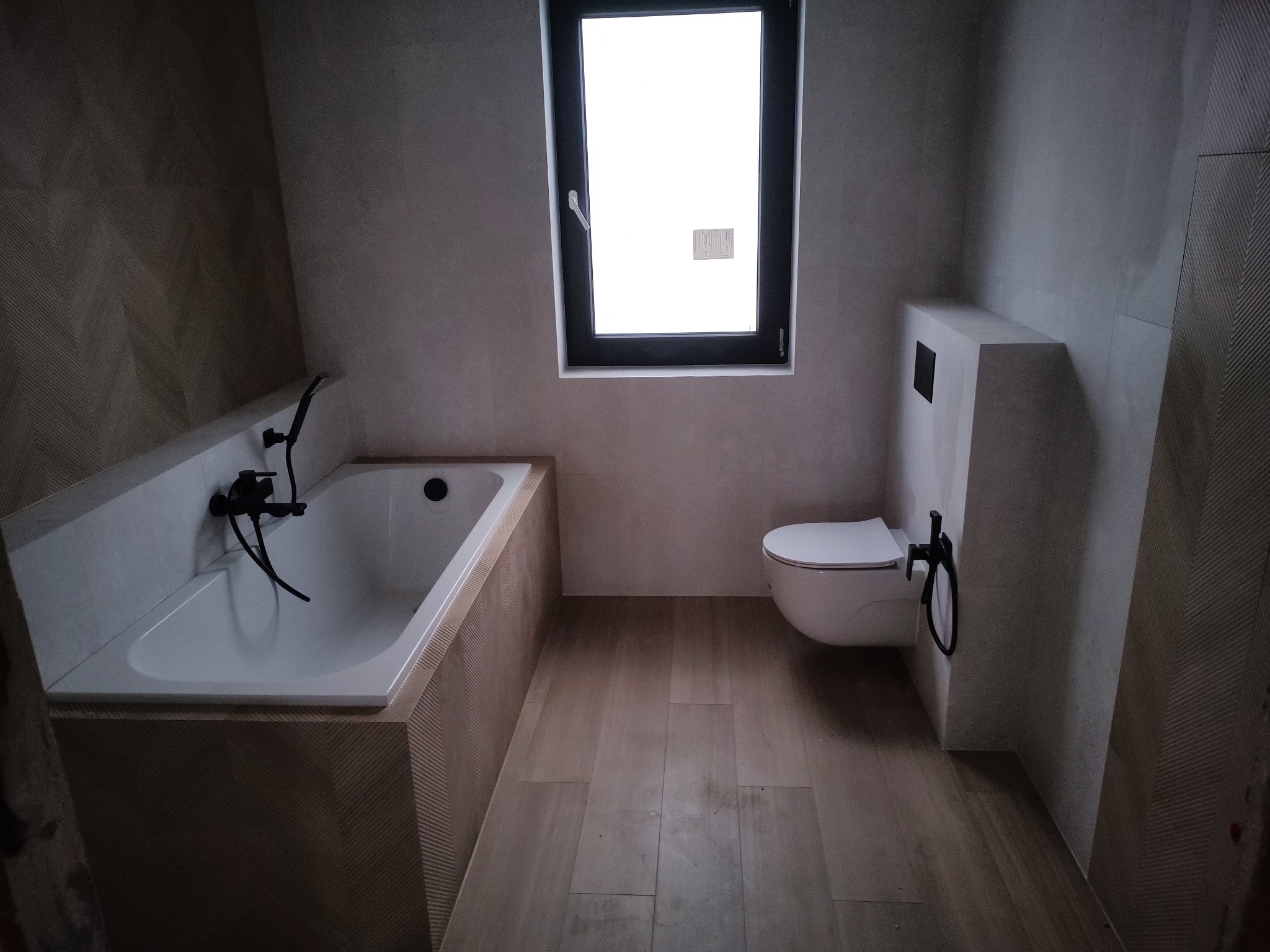 Nowoczesna łazienka z wanną obudowaną płytkami w jodełkę, wiszącą toaletą i czarną armaturą, oświetlona przez okno.