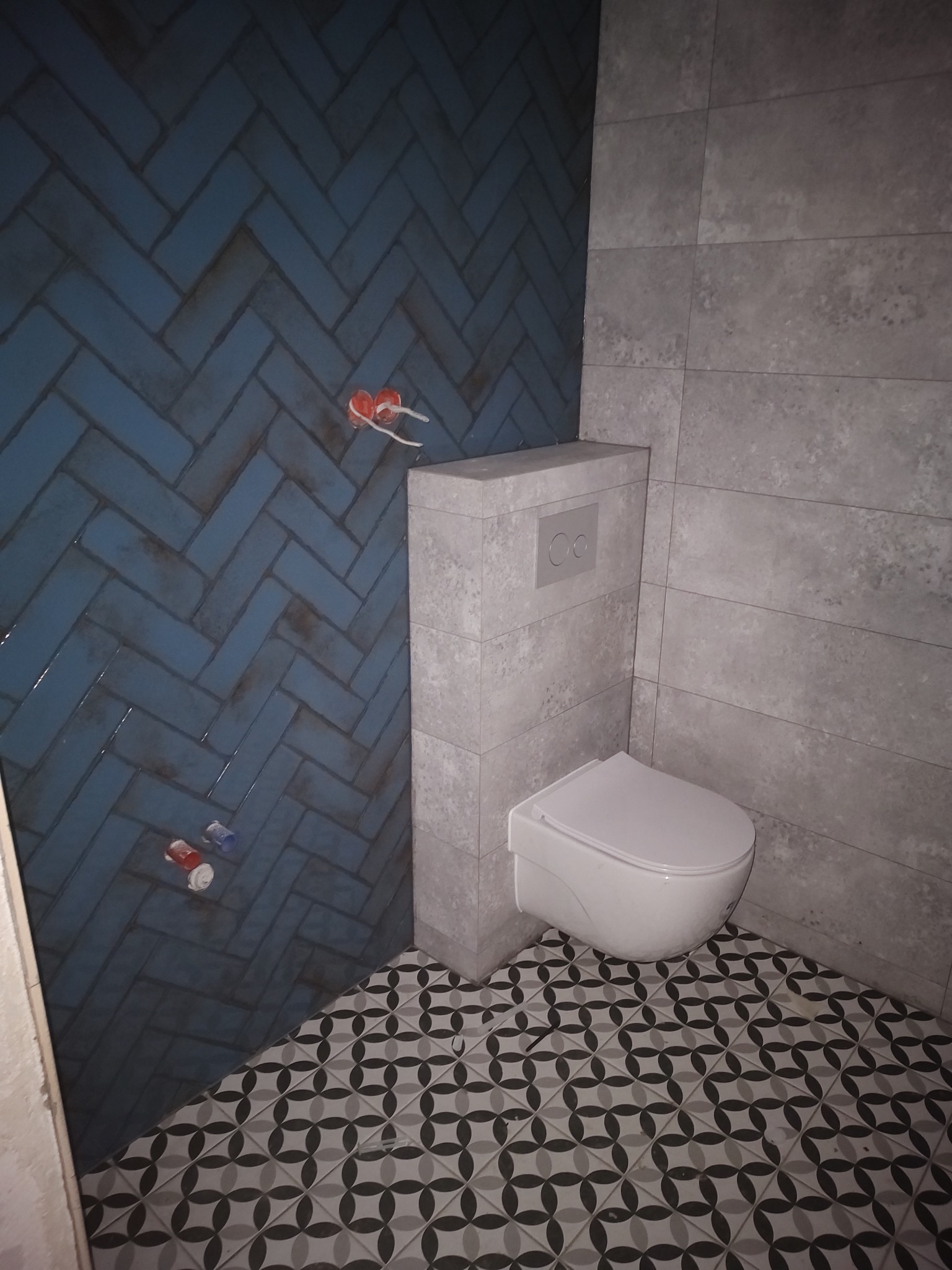 Niebieska ściana z płytek ułożonych w jodełkę w łazience, widoczne przyłącza hydrauliczne, szara ściana z płytek, podwieszana toaleta i podłoga z geometrycznym wzorem.