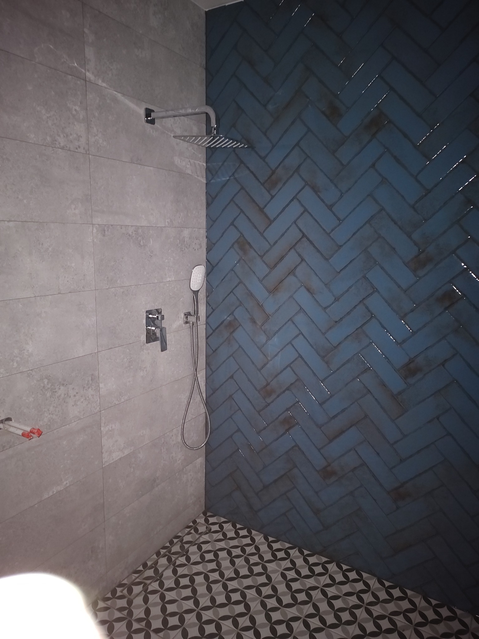 Wykończona kabina prysznicowa z szarymi płytkami imitującymi beton, niebieskimi płytkami ułożonymi w jodełkę i wzorzystą podłogą, widoczne elementy armatury i rury instalacyjne.