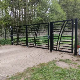 Czarna, metalowa brama wjazdowa z automatyką na posesji, widok z zewnątrz na tle drzew i siatki ogrodzeniowej, kostka brukowa na podjeździe.