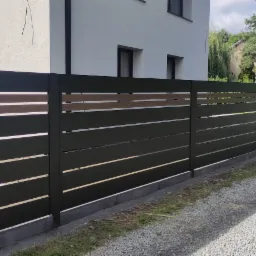 Nowoczesne, poziome przęsła ogrodzeniowe w kolorze grafitowym, z elementami imitującymi drewno, na tle białego domu i żwirowej drogi.