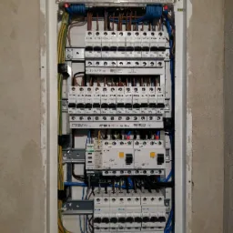 Rozdzielnica elektryczna z widocznymi bezpiecznikami, przewodami w kolorach niebieskim, brązowym, żółto-zielonym, zamontowana w ścianie z surowym wykończeniem, z widocznymi elementami montażowymi.