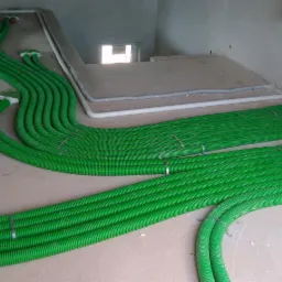 Zielone, karbowane rury wentylacyjne ułożone równolegle na surowej podłodze w budynku w trakcie budowy, widoczne mocowania i fragment instalacji elektrycznej w ścianie.