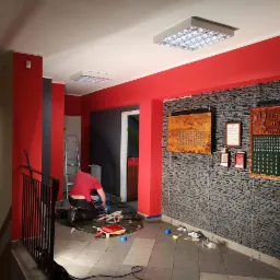 Pracownik w czerwonej koszulce przygotowuje podłoże do położenia podłogi w pomieszczeniu z czerwonymi i szarymi ścianami, widoczne narzędzia i materiały, na ścianie wisi tablica pamiątkowa OSP...