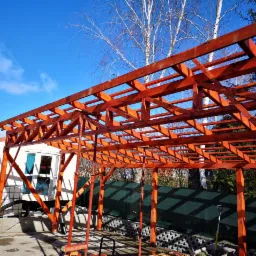 Konstrukcja drewniana z belek, prawdopodobnie zadaszenie tarasu lub wiaty, w trakcie montażu, pomalowana na brązowo, widoczne niebo i drzewa w tle.