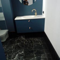 Gres-Bud - Niebieska szafka łazienkowa z białą umywalką i złotą baterią na tle czarnej marmurowej podłogi, widoczne odbicie osoby w okrągłym lustrze.