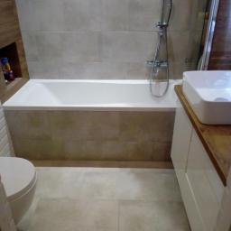 Gres-Bud - Wykończona łazienka z beżowymi płytkami, białą wanną obudowaną płytkami, toaletą i umywalką na szafce z drewnianym blatem. W ścianie wnęka z dekoracjami.