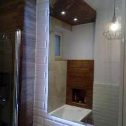 Gres-Bud - Nowoczesna łazienka z podświetlanym lustrem w ramie z białych płytek typu metro, odbijającym drewniany sufit i fragment wanny oraz dekoracyjną lampę wiszącą.