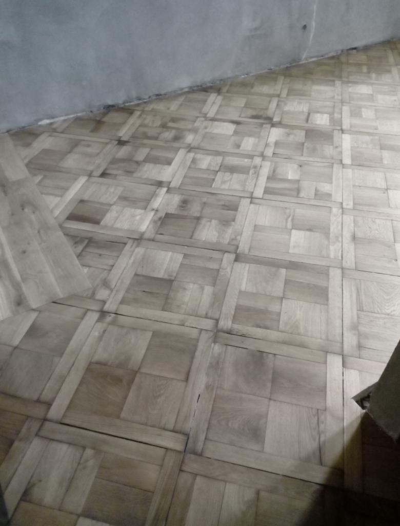 Układanie parkietu w jodełkę francuską z jasnego drewna, widoczny fragment surowej ściany i niedociągnięcia w wykończeniu.