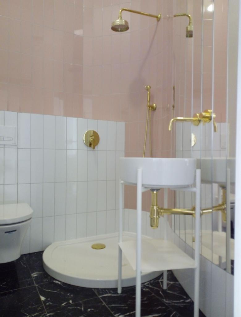 Nowoczesna łazienka z różowymi i białymi płytkami, armatura w kolorze złota, umywalka na białym stelażu, czarna marmurowa podłoga.