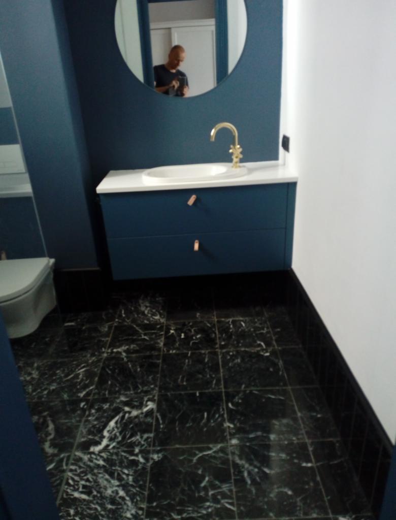 Niebieska szafka łazienkowa z białą umywalką i złotą baterią na tle czarnej marmurowej podłogi, widoczne odbicie osoby w okrągłym lustrze.