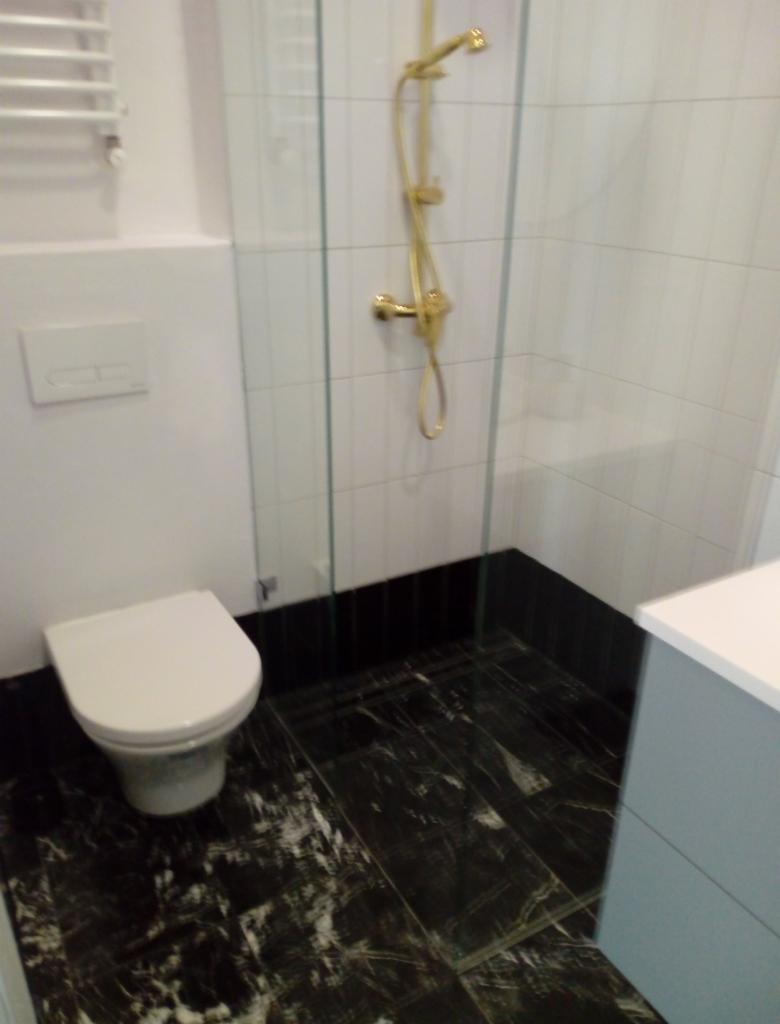 Nowoczesna łazienka z czarną marmurową podłogą, białą ceramiką i złotą armaturą prysznicową za szklaną kabiną.