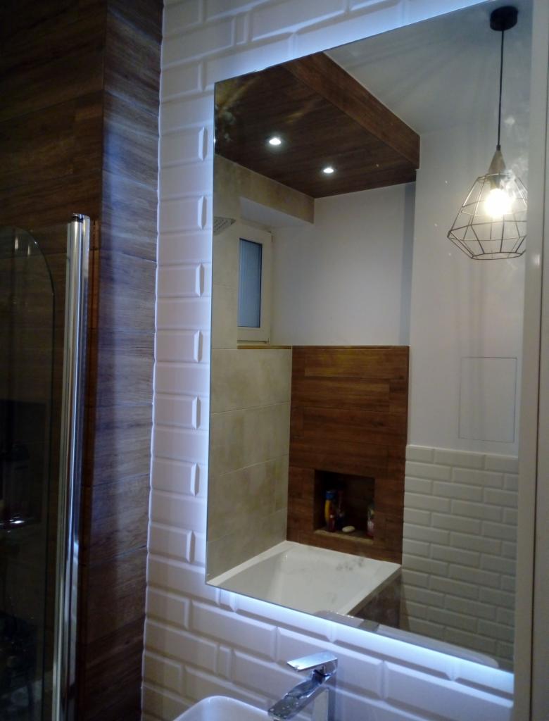 Nowoczesna łazienka z podświetlanym lustrem w ramie z białych płytek typu metro, odbijającym drewniany sufit i fragment wanny oraz dekoracyjną lampę wiszącą.