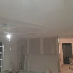 Wnętrze w trakcie remontu: widoczne ściany z płyt kartonowo-gipsowych, sufit z zamontowaną konstrukcją podwieszaną w kształcie trapezu, drabina aluminiowa przy oknie i narzędzia budowlane na podłodze.