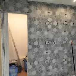 Ściana w trakcie układania szarych, heksagonalnych płytek z wzorem, widoczne krzyżyki dystansowe, poziomica oparta o ścianę w przejściu do innego pomieszczenia, obok stoją worki ze śmieciami.