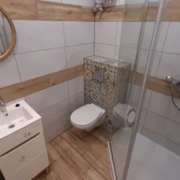 Nowoczesna łazienka z umywalką na szafce, podwieszaną toaletą z zabudową w dekoracyjne płytki i kabiną prysznicową; widoczne lustro owalne z odbiciem osoby robiącej zdjęcie.