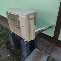 Zewnętrzna jednostka klimatyzacji Atlantic R32 zamontowana na betonowych podstawach przy ścianie budynku o jasnej elewacji, z widoczną rurą odprowadzającą wilgoć owiniętą srebrną folią izolacyjną...