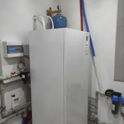 Biały piec grzewczy Atlantic z niebieskim zbiornikiem wyrównawczym i instalacją hydrauliczną w pomieszczeniu technicznym, widoczny filtr wody i rozdzielnia elektryczna na ścianie.