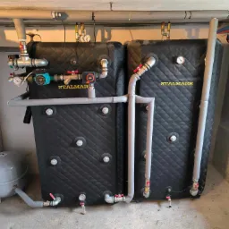 Dwa czarne akumulatory ciepła marki Stalmark z kompletną instalacją hydrauliczną, w tym rury izolowane szarą pianką, zawory z czerwonymi pokrętłami, manometry i pompy obiegowe, umieszczone...