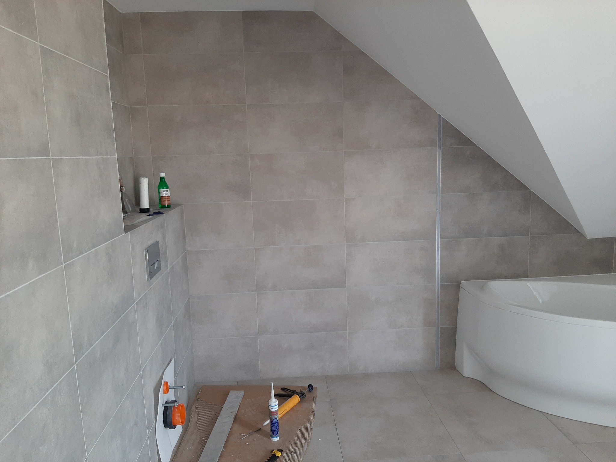 Łazienka na poddaszu w trakcie wykańczania: szare płytki, biała wanna, widoczne narzędzia glazurnicze i elementy instalacji podtynkowej.