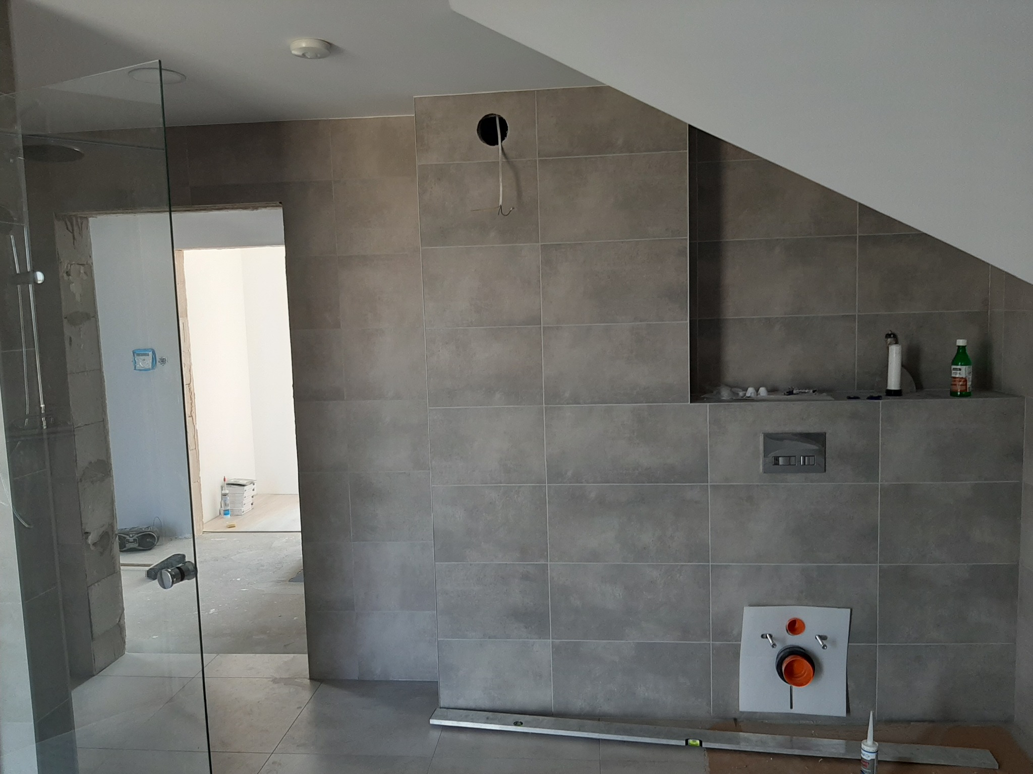 Szare płytki na ścianie łazienki w trakcie montażu armatury, widoczna instalacja podtynkowa WC, poziomica i częściowo zamontowana kabina prysznicowa.