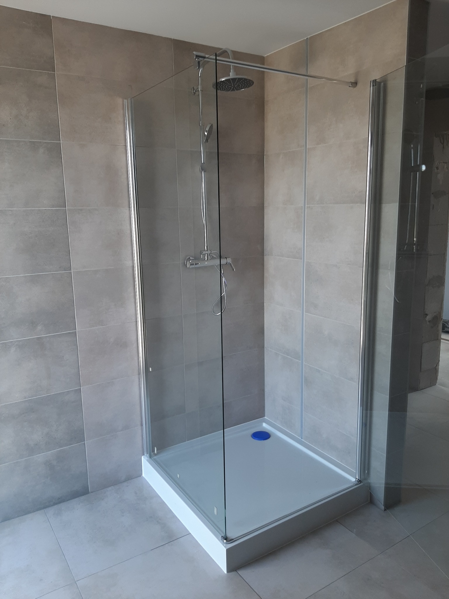 Nowoczesna kabina prysznicowa z chromowanymi elementami, zamontowana w łazience wyłożonej szarymi płytkami ceramicznymi. Widoczny niebieski odpływ w brodziku.