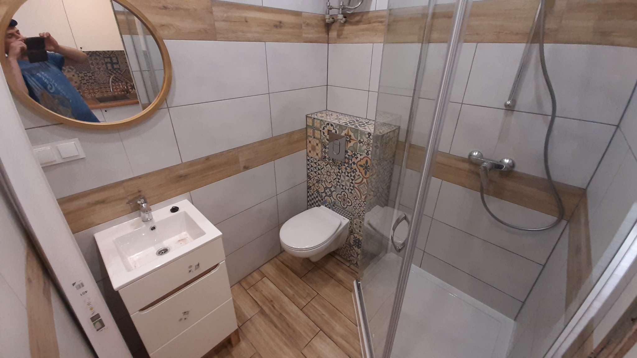 Nowoczesna łazienka z umywalką na szafce, podwieszaną toaletą z zabudową w dekoracyjne płytki i kabiną prysznicową; widoczne lustro owalne z odbiciem osoby robiącej zdjęcie.