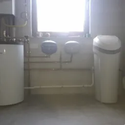 Kotłownia z piecem Vaillant, dwoma zbiornikami wyrównawczymi, zmiękczaczem wody i instalacją hydrauliczną z miedzianych rur, oświetlona lampą na statywie w rogu pomieszczenia.