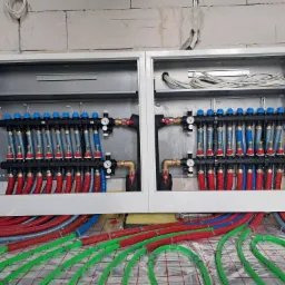 Rozdzielacz ogrzewania podłogowego w szafce, widoczne rury w kolorach czerwonym, niebieskim i zielonym na folii izolacyjnej z siatką, ściany z betonu komórkowego.