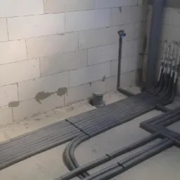 Szare rury grzewcze w izolacji ułożone na podłodze w surowym pomieszczeniu z białymi ścianami z betonu komórkowego, widoczne pionowe wyprowadzenia rur.