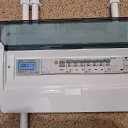 Otwarta skrzynka elektryczna z licznikiem Luno Pro i zabezpieczeniami Eaton, zamontowana na ścianie o wzorzystej strukturze, widoczne białe rury instalacyjne u góry.