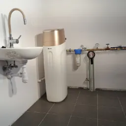 Biała umywalka z elastyczną baterią, obok zmiękczacz wody z elementami w kolorze złotym i instalacja wodna z filtrem i manometrami na tle szarych płytek podłogowych.