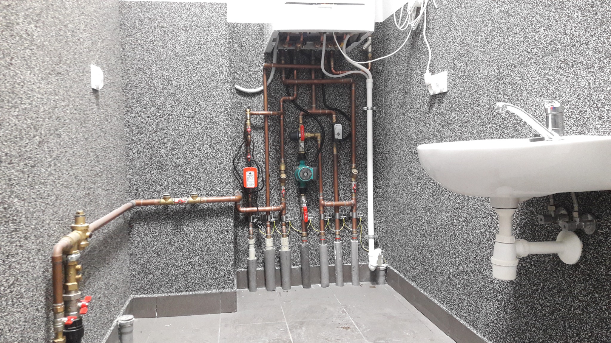 Węzeł cieplny w kotłowni z piecem gazowym, rury miedziane, zawory, pompa obiegowa i umywalka na tle ściany o strukturze baranka.