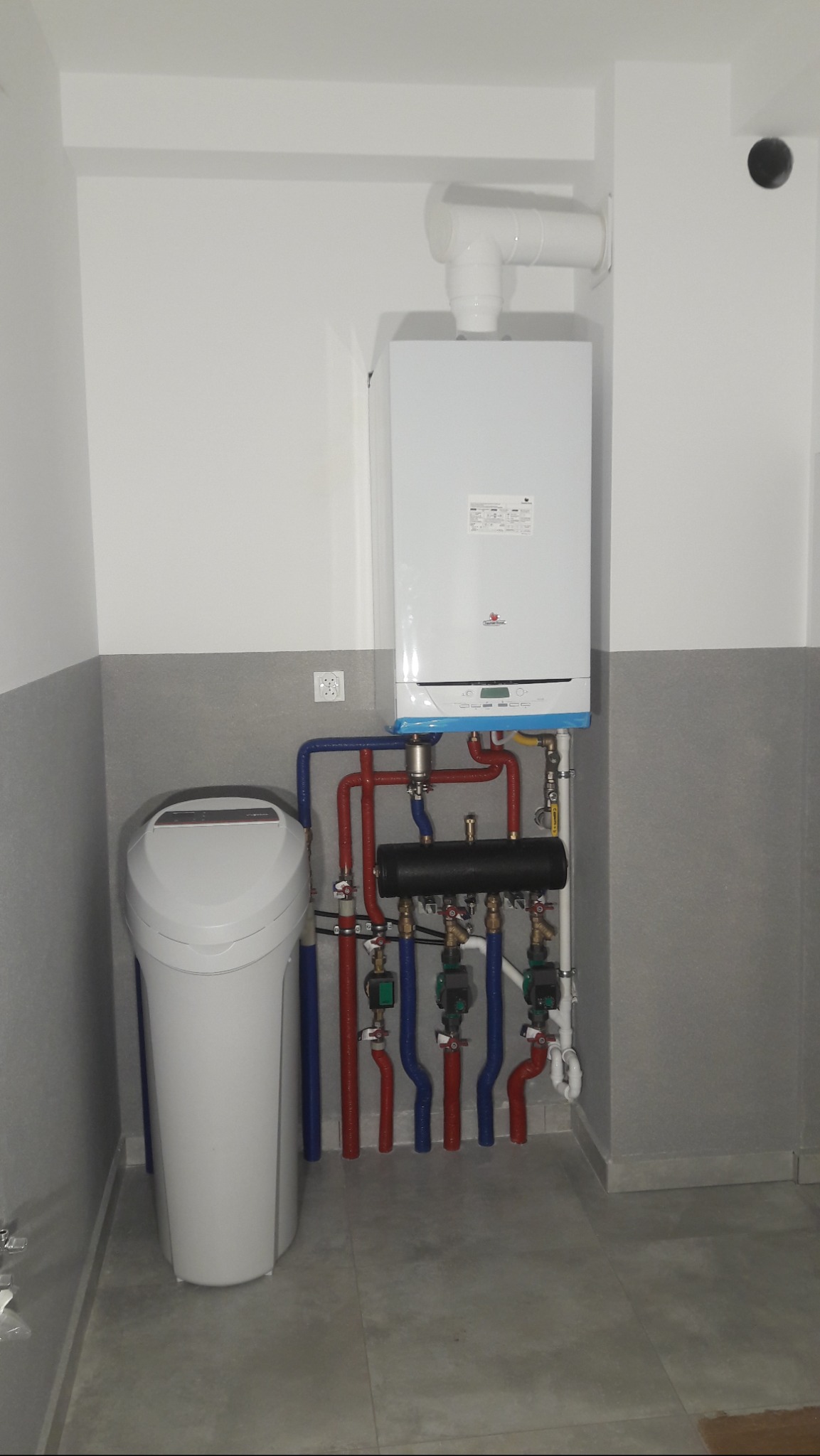Biały piec gazowy zamontowany na ścianie, z podłączonymi rurami w kolorach niebieskim i czerwonym oraz czarnym rozdzielaczem, obok stoi biały zmiękczacz wody.