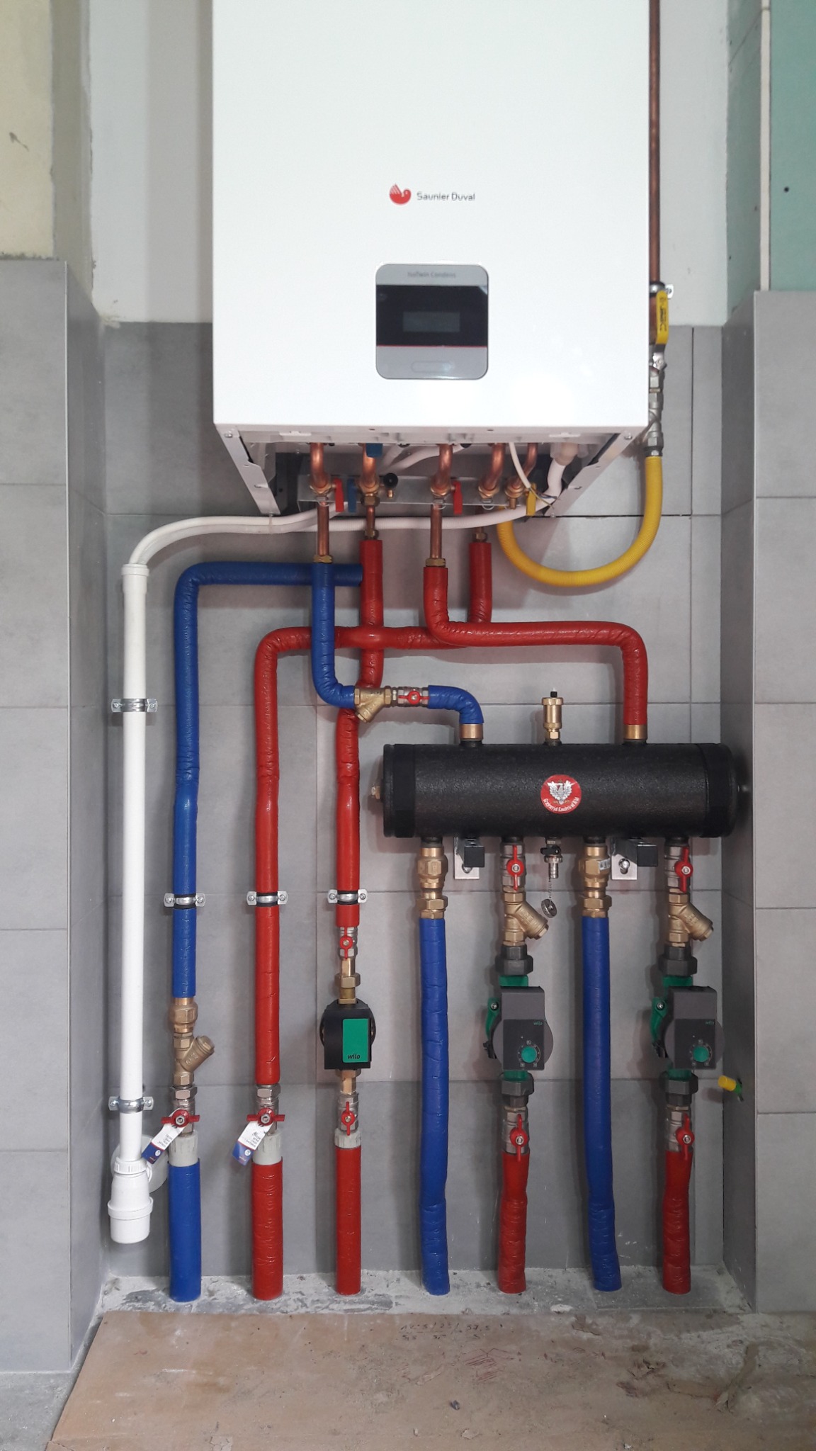 Nowoczesny piec gazowy Saunier Duval z rozdzielaczem i rurami w izolacji czerwonej i niebieskiej, zamontowany na szarej, kafelkowanej ścianie.