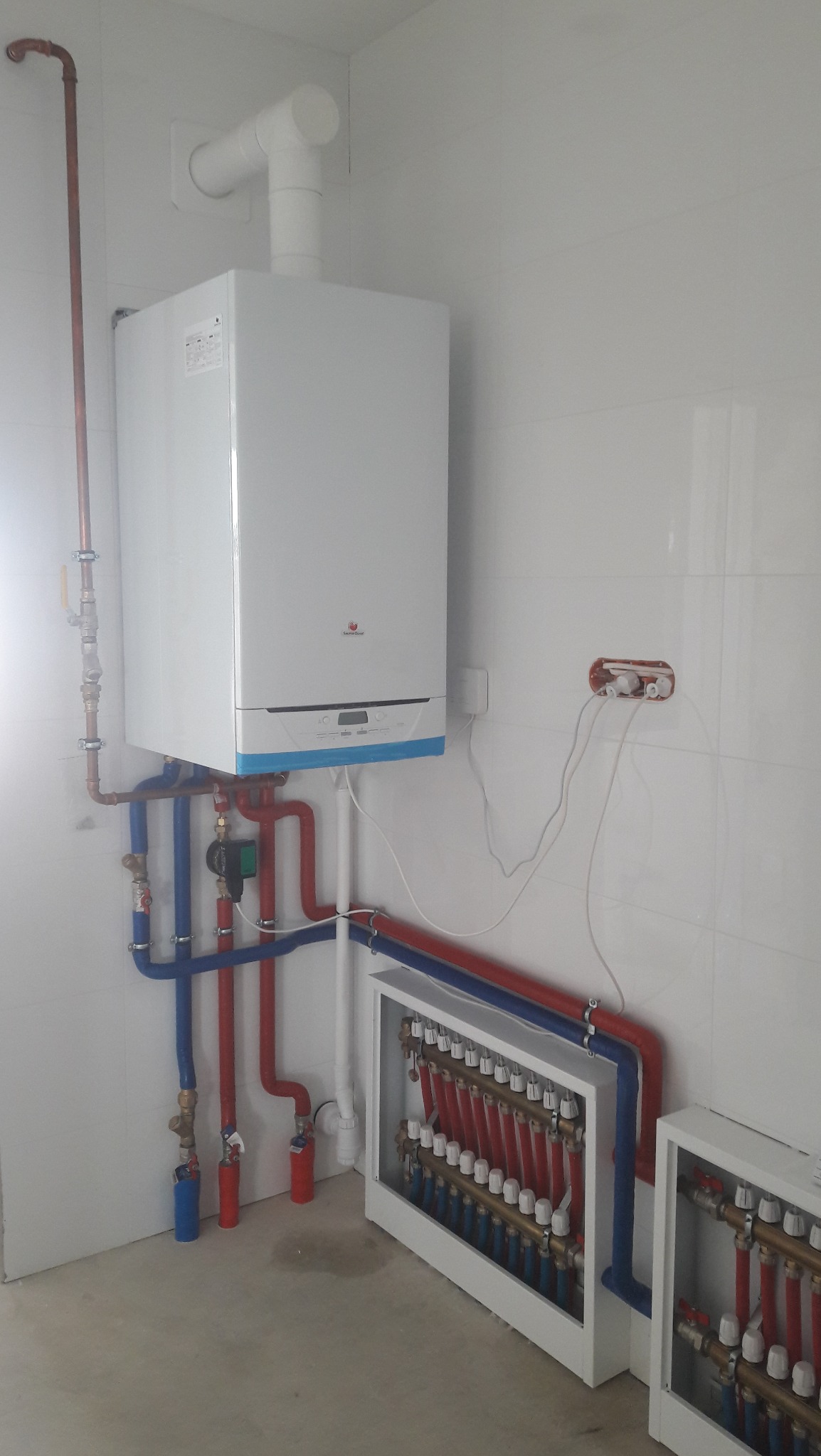 Nowoczesny, wiszący piec gazowy z podłączonymi rurami miedzianymi i plastikowymi (czerwone i niebieskie) oraz rozdzielaczami ogrzewania podłogowego w białej obudowie na tle białych płytek.