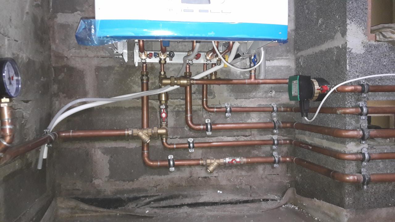 Nowy piec gazowy z niebieską folią ochronną, podłączony do instalacji z miedzianych rur, widoczny manometr i pompa obiegowa, surowa ściana z betonu komórkowego w tle.