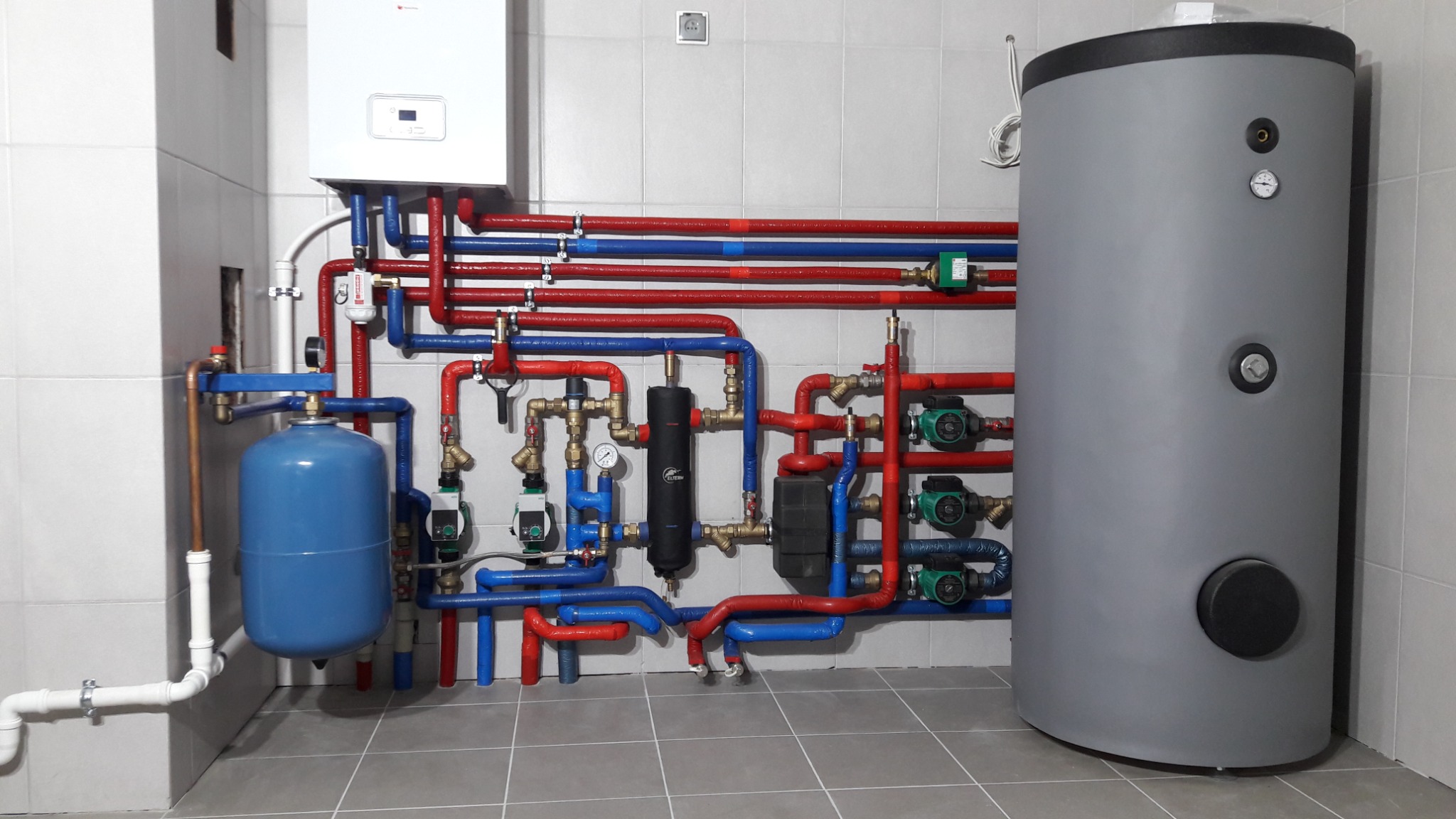 Kotłownia z szarym zasobnikiem ciepłej wody, białym piecem gazowym, niebieskim zbiornikiem wyrównawczym i rurami w izolacji czerwonej i niebieskiej, widok na całą instalację.