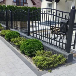 Nowoczesna posesja z szarą kostką brukową, czarnym metalowym ogrodzeniem z numerem 7 na skrzynce pocztowej i równo przyciętymi krzewami, widok z poziomu gruntu.